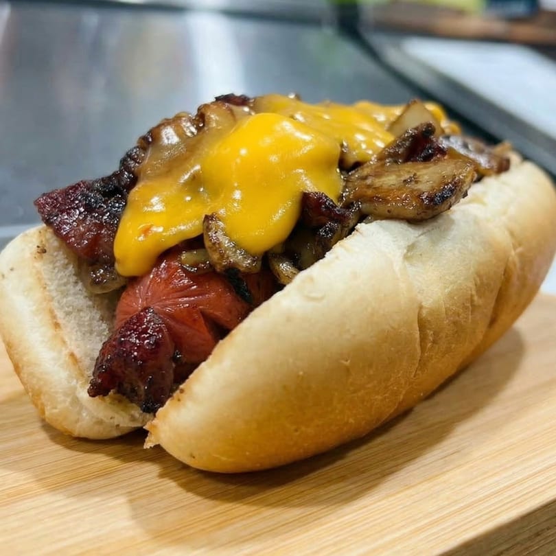 Checkmate Burgers' hot dog with melted cheese, bacon, and mushrooms on a wooden board.