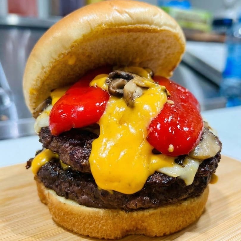 A double Checkmate Burger with melted cheese, roasted red peppers, and mushrooms on a wooden board.